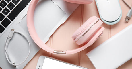 A close-up of modern devices. Pink headphones rest on a laptop keyboard with a mouse, smartphone, and cords around it.