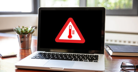 A laptop on a wooden desk with a pen, glasses, a plant, and a notebook nearby. The screen displays a high-temperature warning symbol.