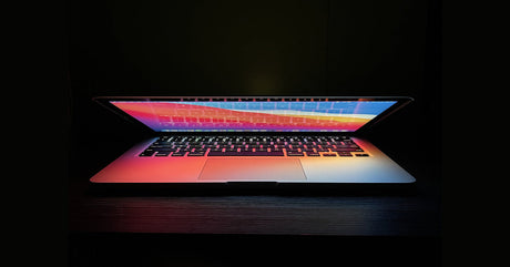 A half-open laptop in a dark room. The display is lit up, casting various colors over the laptop's keyboard.