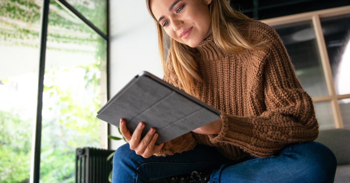 A woman wearing a chunky brown sweater sits on an indoor bench. She holds a tablet in her hands and looks at the screen.