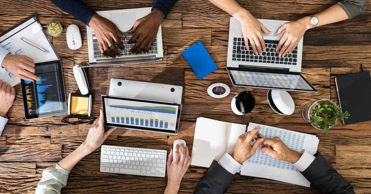 Six people work at a shared wooden table with laptops, tablets, charts, coffee, and more in front of them.