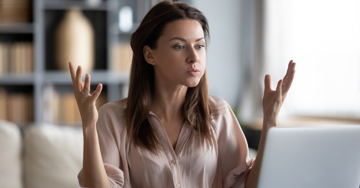 A woman sits in front of a laptop with a notebook open next to it. She is frustrated, with her hands thrown up in front of her.