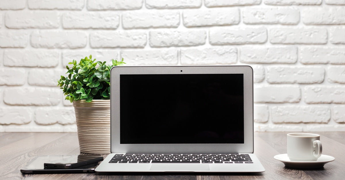 An open laptop with a black screen rest son a wooden table. A phone, a tablet, a coffee cup, and a potted plant rest near the laptop.