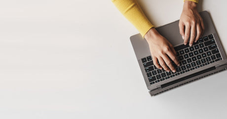 A bird's-eye view of someone with their hands on a laptop keyboard, typing. The laptop rests on a white table.