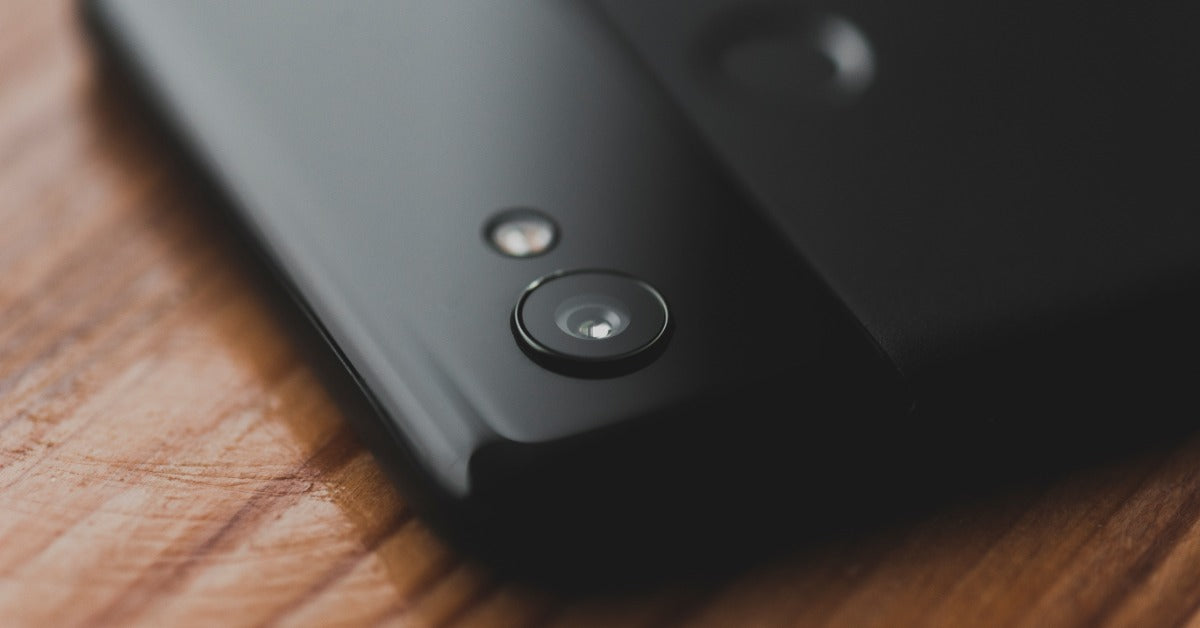 A close-up of the back camera of a black smartphone on a wooden table. The rest of the phone is blurry.