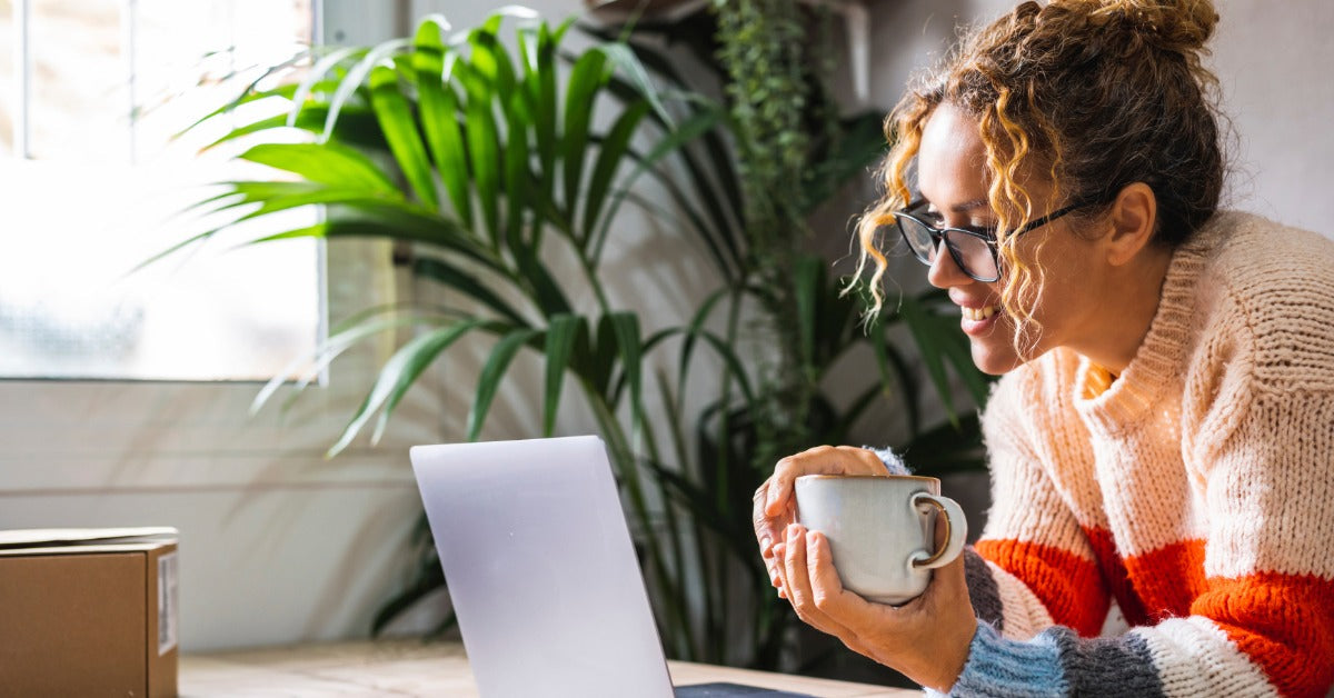 A woman holding a coffee mug leans against a table in front of an open laptop. A large plant is behind the table.